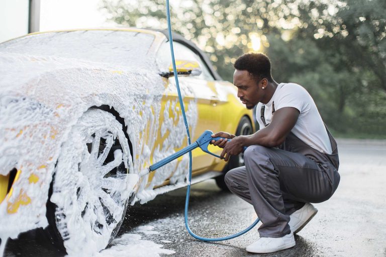 Young African man with high pressure jet spraying the foam on car wheel at car wash service station